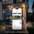 Homeowner using a smart home security app on a smartphone to remotely monitor a front door camera feed, control smart locks, and view real-time activity logs outside a modern house at dusk.