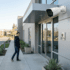 White outdoor bullet security camera mounted on a modern commercial building exterior near a glass entrance, providing surveillance for a passerby walking near a "Protected" security sign.